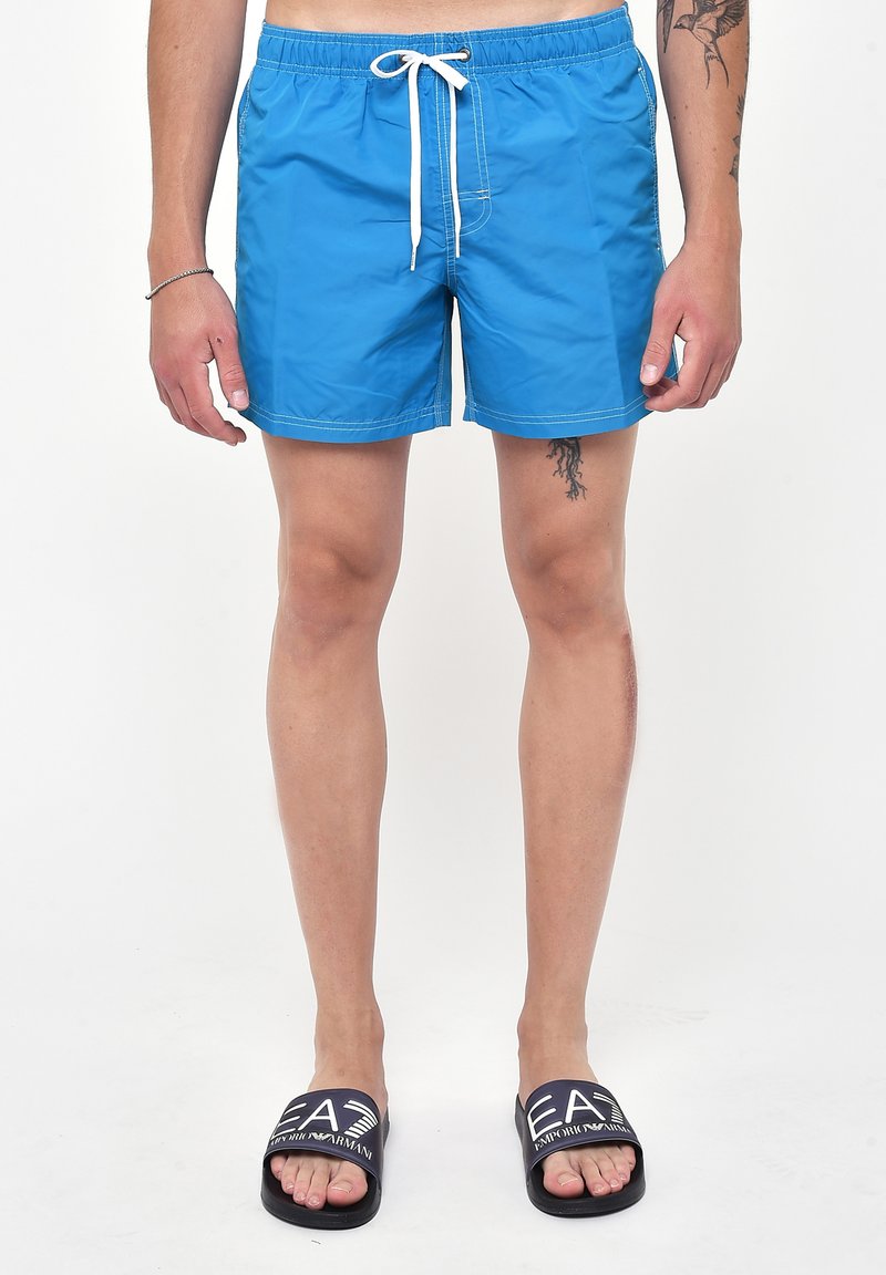 Shorts da bagno blu con cintura elastica, lacci bianchi e cuciture a contrasto; abbinati a sandali neri con logo.