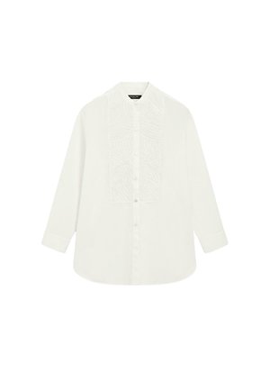 Kobieta stoi przy ścianie, ubrana w białą, teksturowaną bluzkę, szerokie czarne spodnie i czarne sandały na koturnie z paskami na palce.