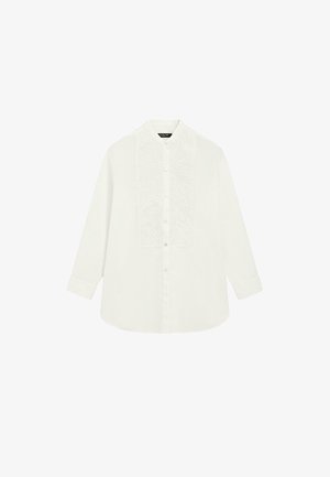 Kobieta stoi przy ścianie, ubrana w białą, teksturowaną bluzkę, szerokie czarne spodnie i czarne sandały na koturnie z paskami na palce.