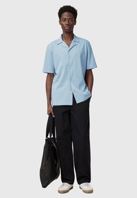 Camicia a maniche corte azzurro chiaro con colletto, bottoni davanti; abbinata a pantaloni neri larghi e sneakers bianche. Tiene una borsa nera.