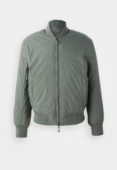 Olivgrön bomberjacka med dragkedja framtill, ribbat krage och manschetter, med en texturerad mönster av upprepade logotyper och två sidfickor.