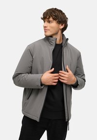 Siva zip-up jakna z visokim ovratnikom, izdelana iz gladkega teksturiranega materiala. Občudovana je nad črno jopico, z vidnimi stranskimi žepi.