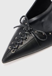 Chaussure en cuir noire à bout pointu avec des lacets de style corset et de petites clous en argent le long du bord supérieur.