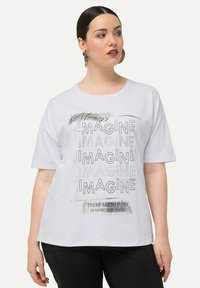 Hvid T-shirt med korte ærmer, der har et grafisk design med det gentagne ord "IMAGINE" og en tekstaccent. Lavet af blødt stof, afslappet pasform.