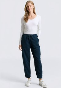 Merimiesmolekyyliset housut, joissa on joustava vyötärönauha ja sivutaskut, yhdistettynä pitkähihaiseen valkoiseen paitaan ja valkoisiin lenkkareihin.