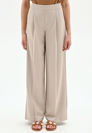 Pantaloni beige a gamba larga con sottili righe, indossati con sandali piatti marroni decorati con grandi gemme trasparenti.