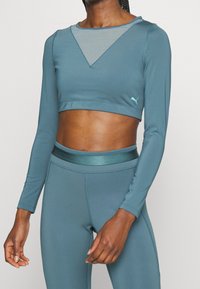 Long-sleeve teal crop top with a v-neck design and contrasting mesh panel. Features a fitted waist and a small logo accent.
