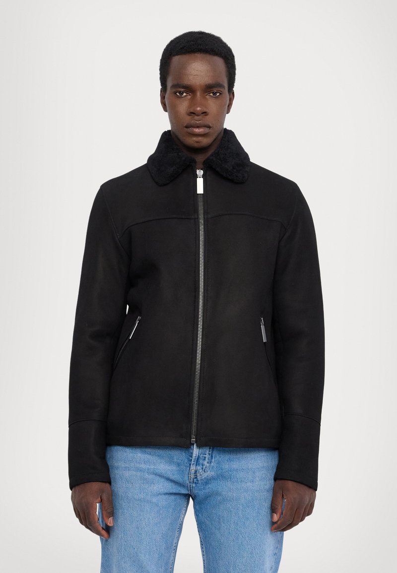 Schwarze Kunstlederjacke mit einem weichen schwarzen Fellkragen, Reißverschluss und zwei Seitentaschen, getragen über blauen Jeans.
