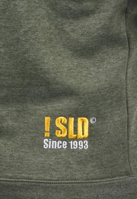 Olive green sweatshirt with a soft texture featuring embroidered text: "! SLD" in yellow and "Since 1993" in white. Relaxed fit design.