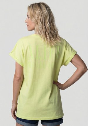 Frau mit welligen blonden Haaren, die ein locker sitzendes, blassgelbes T-Shirt und hochgekrempelte dunkle Jeansshorts trägt, steht mit einer Hand in der Hüfte.