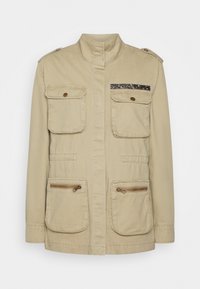 Beige, lehká bunda s vysokým límcem, několika předními kapsami a dekorativním tmavým akcentem na hrudi. Hladký materiál.