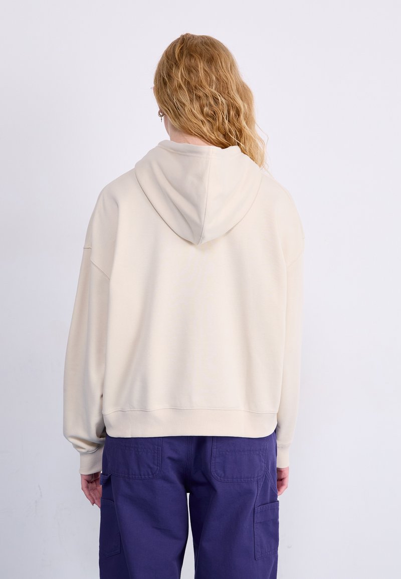 Cremefarbener Kapuzenpullover mit lässigem Schnitt, geripptem Saum und weicher Textur. Kombiniert mit dunkelvioletten Cargo-Hosen mit Seitentaschen.
