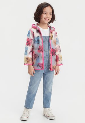 Bambino sorridente, indossa un impermeabile colorato con stampa di Topolino e bordi rosa, maglietta bianca, salopette di jeans e scarpe da ginnastica bianche, in piedi su sfondo bianco.