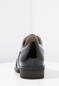 Chaussure en cuir verni noir avec un petit talon, présentant des décorations perforées et une finition lisse et brillante. L'arrière a une forme arrondie.