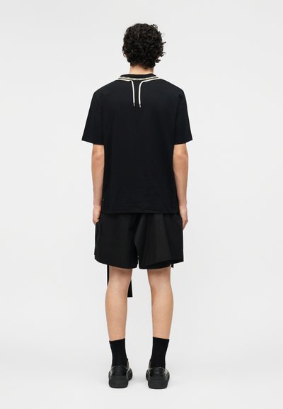 Άνδρας με σγουρά μαλλιά που φοράει μαύρο κοντομάνικο πουκάμισο, άνετα μαύρα σορτς, μαύρες κάλτσες και μαύρα παπούτσια, όρθιος με την πλάτη προς τα πίσω σε απλό φόντο.