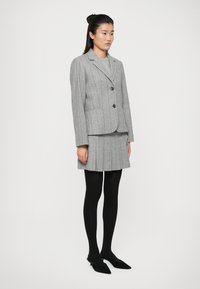 Mulher em pé, vestindo um blazer cinza risca de giz, saia plissada a condizer, meias pretas e sapatos de salto agulha pretos, contra um fundo branco.