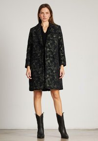 Mujer vestida con un abrigo negro de estampado floral hasta la rodilla sobre un vestido negro y botas de cuero negras hasta la mitad de la pantorrilla, de pie contra un fondo liso.