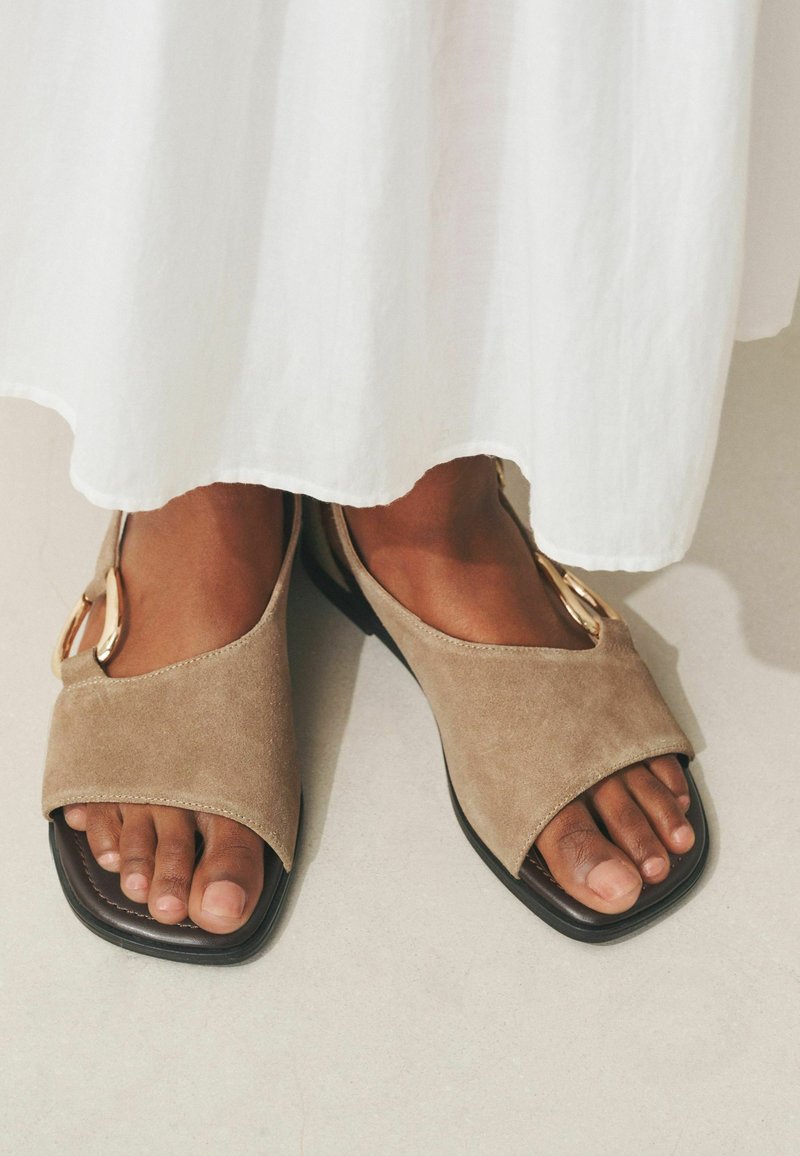 Pieds portant des sandales ouvertes à bout ouvert en daim beige avec un détail de boucle dorée, associés à un vêtement blanc fluide, sur un sol clair.