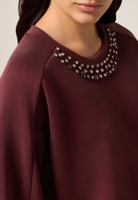 Bordeaux top met een gladde textuur, met een ronde halslijn versierd met heldere en donkerrode rhinestones.
