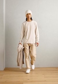 Beige hosszúujjú ing és hozzá illő bézs nadrág egy kötött bézs sapkával. Egy világos csíkos takarót tart, fa padlón áll.