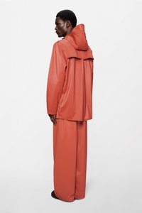 Αδιάβροχο πορτοκαλί μπουφάν βροχής με κουκούλα, μακριά μανίκια και πίσω πτερύγιο, σε συνδυασμό με αντίστοιχο φαρδύ παντελόνι. Λεία υφή και μινιμαλιστικός σχεδιασμός.