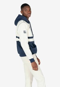 Hætte sweatshirt i navy og creme farver, med en frontlomme, vandrette striber og logo detajle på ærmet. Blød stoftekstur.