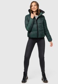 Green puffer jacket with a high collar, zip closure, and black accents. Model wears black jeans and lace-up boots. Smooth texture.