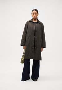 Woman wearing a dark oversized coat, wide-leg jeans, black shoes, and hoop earrings, holding an olive green handbag against a plain background.