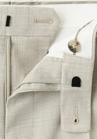 Gros plan d’un pantalon beige à motif pied-de-poule montrant la ceinture, le bouton, la boutonnière, la fermeture éclair et la doublure intérieure blanche en tissu.
