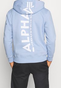 Felpa leggera blu con un grande logo bianco "ALPHA" sul retro. Presenta un cappuccio con lacci, polsini a coste e una tasca con zip sulla manica.