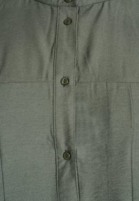 Camisa verde oscura de tela suave, con cierre de botones en el frente, mangas cortas y dos paneles horizontales cosidos.