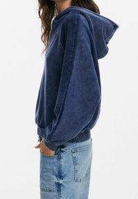 Hoodie blu navy realizzato in tessuto morbido, dotato di cappuccio con cordino, vestibilità ampia e polsini a coste. Abbinato a jeans in denim chiaro.