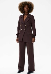 Bruine dubbele blazer met gouden knopen, zijzakken en lange mouwen, gecombineerd met bijpassende wijde broek en slanke zwarte schoenen.