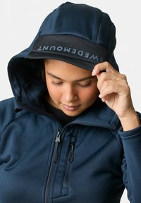 Navyblauwe hoodie met een zwarte klep en logo. Heeft een ritssluiting en de textuur van de stof is zichtbaar rond de schouders en de kraag.