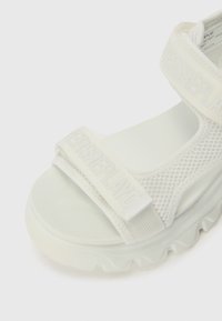 Sandal branco com uma tira de malha texturizada com logótipo em relevo, sola almofadada com um padrão de onda para maior aderência e conforto.