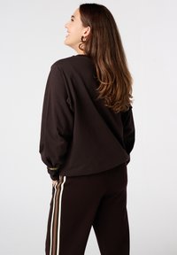 Vrouw met lang bruin haar die glimlacht, gekleed in een donkerbruine sweatshirt en bijpassende broek met witte en bruine zijstrepen, staande tegen een witte achtergrond.