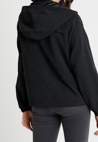 Giacca nera con cappuccio in finitura opaca, spalle cadenti e polsini a coste, caratterizzata da una vestibilità semplice e rilassata e dettagli con apertura sul retro.