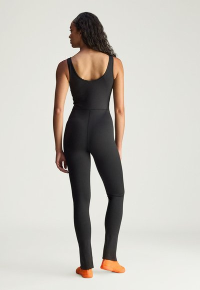 Black fitted jumpsuit with a scoop back, long legs, and side slits, paired with bright orange footwear, showcasing a smooth texture.