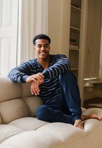 Blauwgrijs gestreept pyjamapak gedragen door een persoon die op een lichtbeige bank zit, met zachte bekleding en natuurlijk licht.