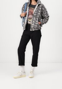 Kurtka polarowa z czarno-białym kwiatowym wzorem, szara koszulka z nadrukiem, czarne spodnie i beżowe trampki. Akcesoria obejmują niebieską torbę.