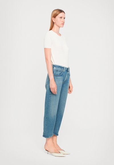 Camicia corta a maniche corte beige chiaro indossata con jeans a vita alta blu medio. I jeans hanno una vestibilità rilassata e un orlo corto. Scarpe bianche slip-on.