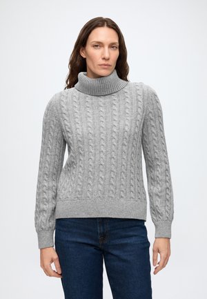 Femme aux longs cheveux bruns portant un pull à col roulé gris en maille torsadée et un jean bleu, debout sur un fond blanc uni.