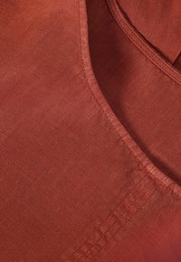 Tejido de color óxido con una textura suave, que presenta un escote curvado y costuras planas. El material parece ligero y transpirable.
