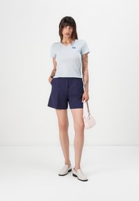Ljusblå V-ringad t-shirt, marinblå shorts och vita slip-on skor. Håller en liten rosa handväska. Minimalistisk design, avslappnad stil.