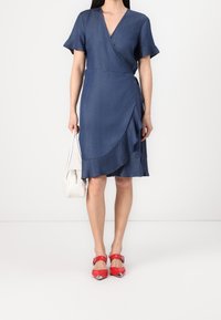Denim wrap dress with short sleeves, V-neck, and ruffled hem. Paired with red pointed-toe heels and a white handbag.