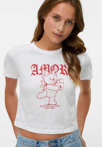 Hvid cropped t-shirt med en rød grafisk dekoration af en teddybjørn med en lås og bånd, samt "AMOR" tekst ovenfor i fed, vintage-stil.