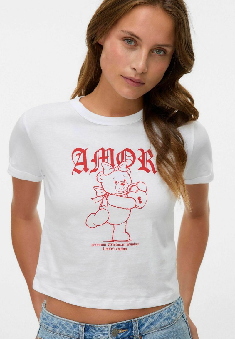 Hvid cropped t-shirt med en rød grafisk dekoration af en teddybjørn med en lås og bånd, samt "AMOR" tekst ovenfor i fed, vintage-stil.