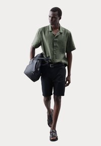 Groene linnen korte mouwen shirt, zwarte shorts, zwarte sandalen. Model draagt een zwarte leren tas met banden. Eenvoudig, gestructureerd ontwerp.