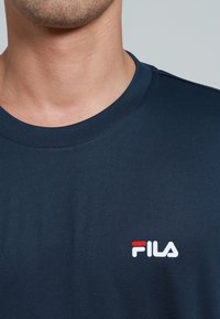 Tmavě modré tričko s krátkými rukávy a kulatým výstřihem, s bílým logem Fila zvýrazněným červenou barvou. Vyrobeno z texturované látky.
