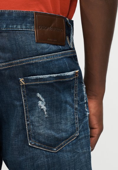 Κοντινή λήψη της πίσω τσέπης σκούρων μπλε φθαρμένων τζιν με καφέ δερμάτινο έμβλημα της μάρκας DSQUARED2 και μέρος του βραχίονα ενός ατόμου.